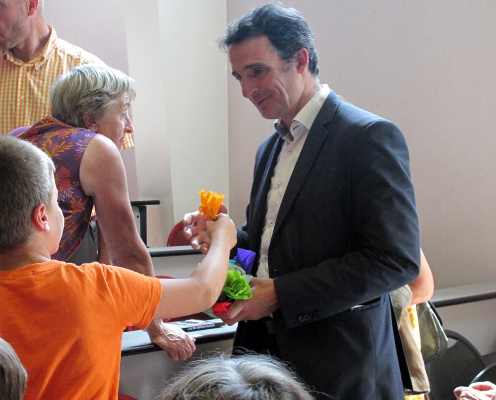 Eric Piolle, maire de Grenoble, recevant des fleurs de papier, à la fin du spectacle donné par les enfants de l'école primaire Bajatière autour du projet "capsule temporelle". Amphithéâtre de l'ESPE. © Séverine Cattiaux – Place Gre’net 